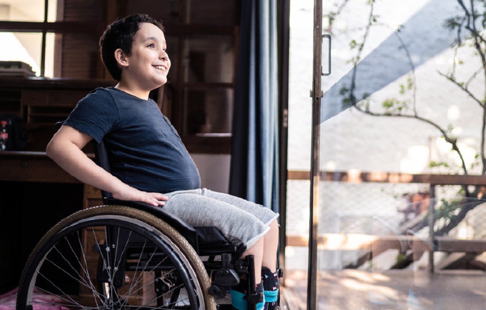Photo d'illustration : Un jeune garçon, assis dans un fauteuil roulant, regarde par la fenêtre avec le sourire. Autonomie. Parcours santé. Parcours patient. Santé.