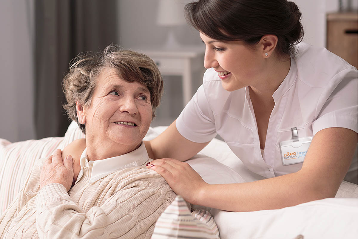 Photo d'illustration Axéo Services : Une infirmière à domicile aux côtés d'une patiente dans le couloir. Elle est souriante. Soins à domicile, Parcours santé. Parcours de soin. Autonomie